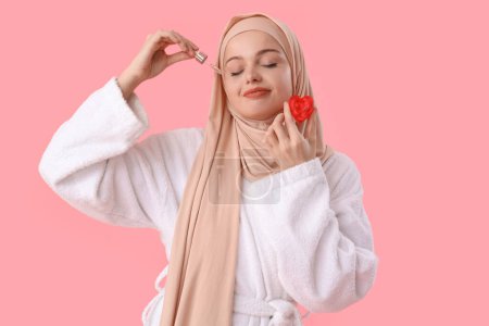 Beautiful Muslim woman in hijab with heart-shaped soap applying serum on pink background