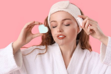 Beautiful woman in bath headband with heart-shaped sponge and makeup foundation on pink background, closeup