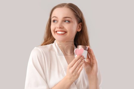 Beautiful young woman with heart-shaped sponge and cream on light background, closeup