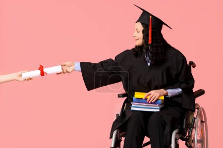 Beautiful female graduate student in wheelchair with graduation cap and hand with diploma on pink background