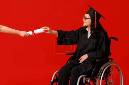 Beautiful female graduate student in wheelchair with graduation cap and hand with diploma on red background