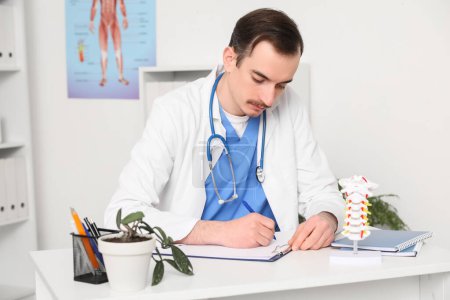 Médico varón escribiendo diagnóstico en la mesa con modelo de columna vertebral en la clínica