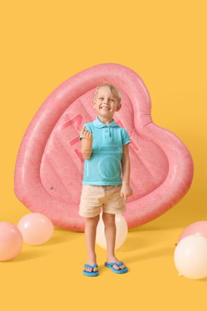 Cute little boy with swim mattress and balloons making heart gesture on yellow background