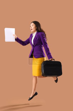 Foto de Divertida empresaria asiática con maletín y documentos saltando sobre fondo marrón - Imagen libre de derechos