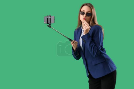 Foto de Divertida empresaria aplicando lápiz labial y tomando selfie sobre fondo verde - Imagen libre de derechos