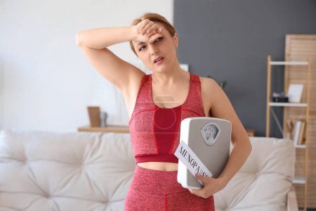 Deportiva mujer madura sosteniendo papel con palabra MENOPAUSE y básculas de peso en casa