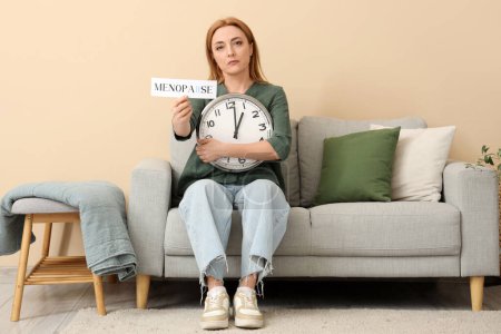 Mature woman holding paper with word MENOPAUSE and clock on sofa at home