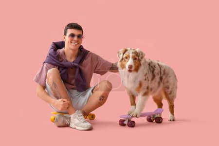 Foto de Joven hombre fresco con perro pastor australiano y monopatines sobre fondo rosa - Imagen libre de derechos