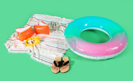 Towel with inflatable ring for swimming, flip flops and headphones on green background. Summer holiday composition