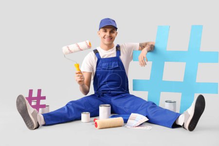 Foto de Pintor masculino con rodillos, latas de pintura y rótulos de hashtag sentado sobre fondo gris - Imagen libre de derechos