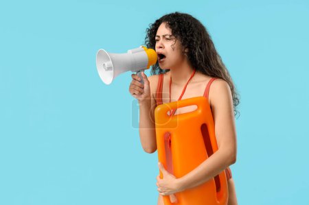 Beautiful young African-American female lifeguard with rescue buoy and megaphone shouting on blue background