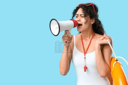 Beautiful young African-American female lifeguard with ring buoy and megaphone shouting on blue background