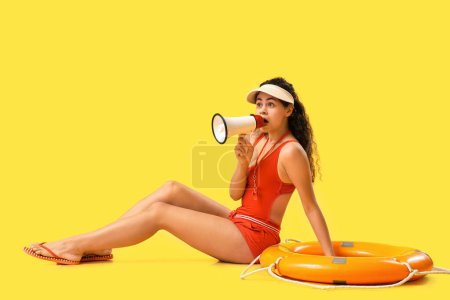 Beautiful young shocked African-American female lifeguard with ring buoy and megaphone on yellow background