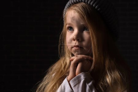 Obdachloses kleines Mädchen betet auf dunklem Hintergrund, Nahaufnahme