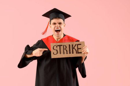 Protesting male graduate pointing at placard with word STRIKE on pink background