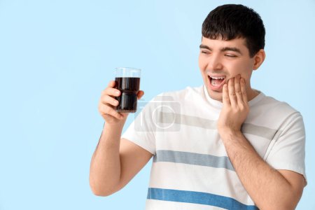 Young man with glass of soda suffering from toothache on blue background, closeup