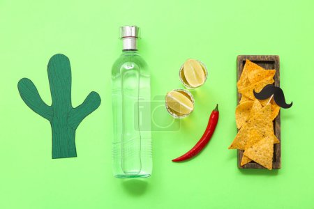 Paper cactus, wooden tray with nachos, chili pepper, bottle and shots of tequila on green background