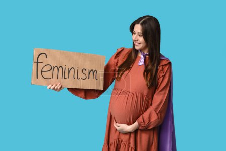 Téléchargez les photos : Belle jeune femme enceinte en cape de super-héros tenant du carton avec le mot FEMINISME sur fond bleu - en image libre de droit