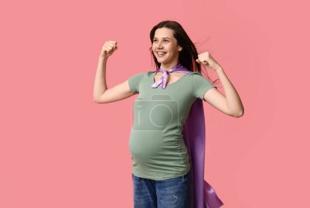 Schöne junge schwangere Frau in Superhelden-Umhang zeigt Muskeln auf rosa Hintergrund