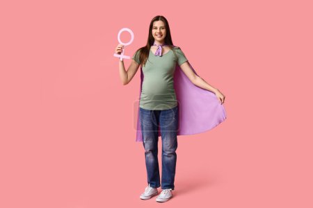 Téléchargez les photos : Belle jeune femme enceinte en cape de super-héros avec symbole de genre sur fond rose - en image libre de droit