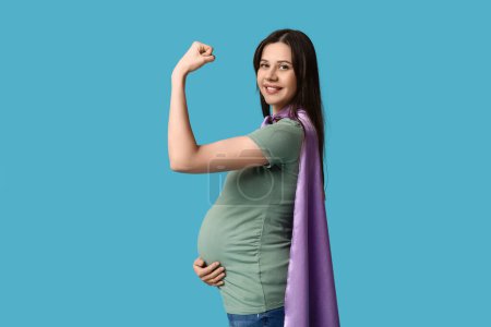 Schöne junge schwangere Frau in Superhelden-Umhang zeigt Muskeln auf blauem Hintergrund