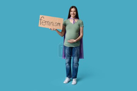 Téléchargez les photos : Belle jeune femme enceinte en cape de super-héros tenant du carton avec le mot FEMINISME sur fond bleu - en image libre de droit