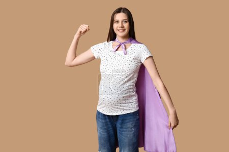 Téléchargez les photos : Belle jeune femme enceinte en cape de super-héros montrant les muscles sur fond brun - en image libre de droit