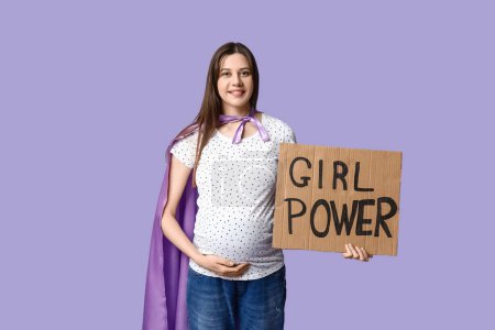 Téléchargez les photos : Belle jeune femme enceinte en cape de super-héros tenant du carton avec texte GIRL POWER sur fond lilas - en image libre de droit