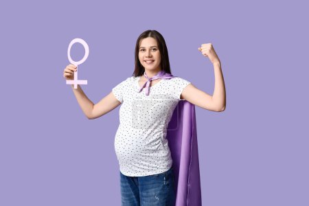 Téléchargez les photos : Belle jeune femme enceinte en cape de super-héros avec symbole de genre montrant les muscles sur fond lilas - en image libre de droit