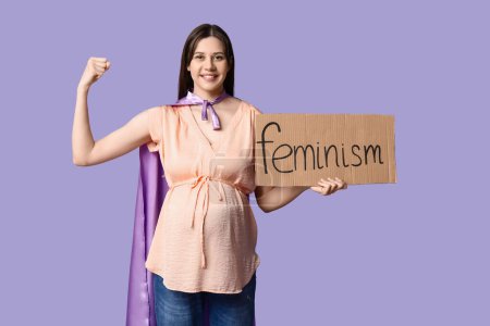 Téléchargez les photos : Belle jeune femme enceinte en cape de super-héros tenant du carton avec le mot FEMINISME et montrant les muscles sur fond lilas - en image libre de droit