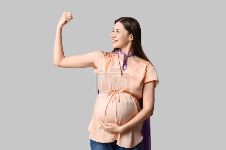 Téléchargez les photos : Belle jeune femme enceinte en cape de super héros montrant les muscles sur fond gris - en image libre de droit
