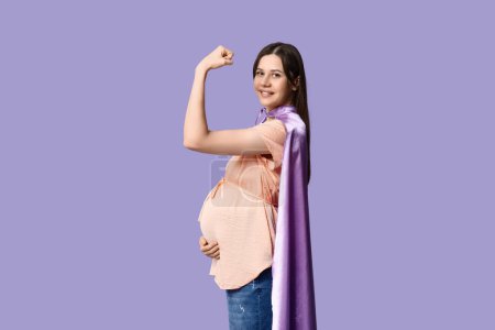 Téléchargez les photos : Belle jeune femme enceinte en cape de super-héros montrant les muscles sur fond lilas - en image libre de droit