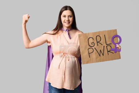 Hermosa joven embarazada en capa de superhéroe sosteniendo cartón con texto GRL PWR y mostrando músculos sobre fondo gris