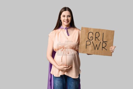 Hermosa joven embarazada en capa de superhéroe sosteniendo cartón con texto GRL PWR sobre fondo gris