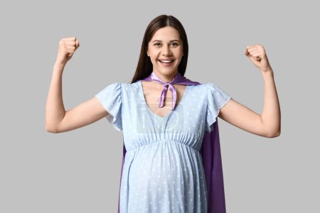 Téléchargez les photos : Belle jeune femme enceinte en cape de super héros montrant les muscles sur fond gris - en image libre de droit