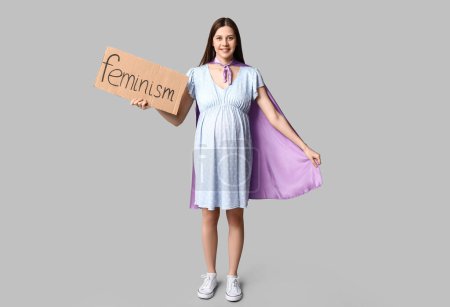 Téléchargez les photos : Belle jeune femme enceinte en cape de super-héros tenant du carton avec le mot FEMINISME sur fond gris - en image libre de droit