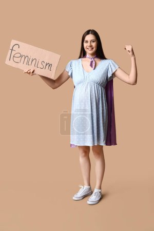 Téléchargez les photos : Belle jeune femme enceinte en cape de super-héros tenant du carton avec le mot FEMINISME et montrant les muscles sur fond brun - en image libre de droit
