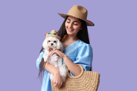 Joven mujer feliz en sombrero con bolsa trenzada y su lindo perro boloñés sobre fondo lila
