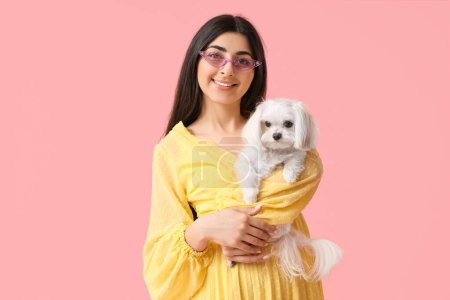 Joven mujer feliz en gafas de sol sosteniendo su lindo perro boloñés sobre fondo rosa