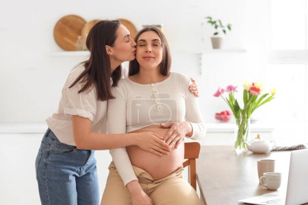 Young lesbian woman kissing her pregnant wife in kitchen