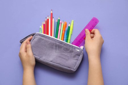 Foto de Manos de niño con estuche de lápiz y diferentes artículos de papelería de la escuela sobre fondo morado - Imagen libre de derechos