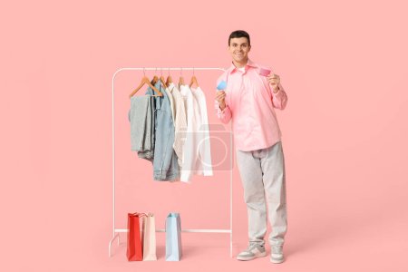 Téléchargez les photos : Beau jeune homme heureux tenant des cartes de crédit et rack avec des vêtements élégants sur fond rose - en image libre de droit