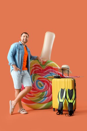 Téléchargez les photos : Jeune homme avec matelas gonflable en forme de bonbons, valise et accessoires de plage sur fond orange - en image libre de droit