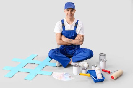 Foto de Pintor masculino con rodillos, latas de pintura y rótulo de hashtag sentado sobre fondo gris - Imagen libre de derechos