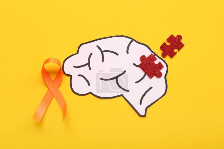 Paper brain with puzzle pieces and orange ribbon on yellow background. Multiple Sclerosis Awareness Month