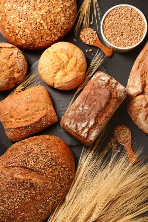 Photo for Loaves of fresh bread with buns, wheat spikelets and grains on black background - Royalty Free Image