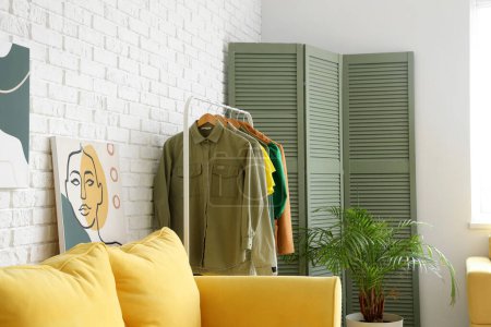 Téléchargez les photos : Support avec vêtements féminins, plante d'intérieur et écran pliant près du mur de briques claires dans la chambre - en image libre de droit