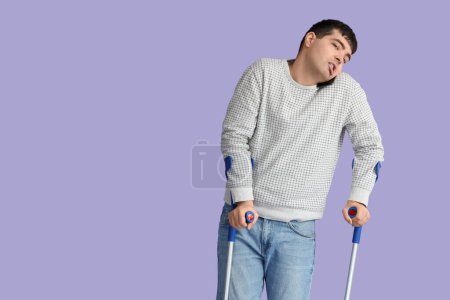 Young man with crutches talking by mobile phone on lilac background. National Cerebral Palsy Awareness Month