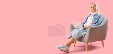 Téléchargez les photos : Programmeur femelle mature avec ordinateur portable dans un fauteuil sur fond rose avec espace pour le texte - en image libre de droit