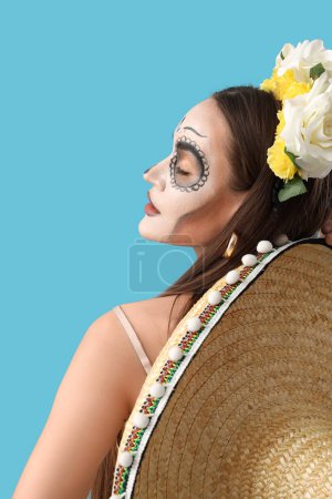 Mujer joven con el cráneo pintado en su cara y sombrero sombrero contra fondo azul claro. Celebración del Día de Muertos de México (El Día de Muertos))